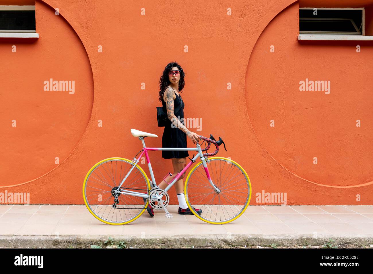 Seitenansicht einer jungen hispanischen Frau mit Sonnenbrille, die in die Kamera schaut, während sie den Griff des Fahrrads hält und auf dem Straßensteg neben der orangefarbenen Wand läuft Stockfoto