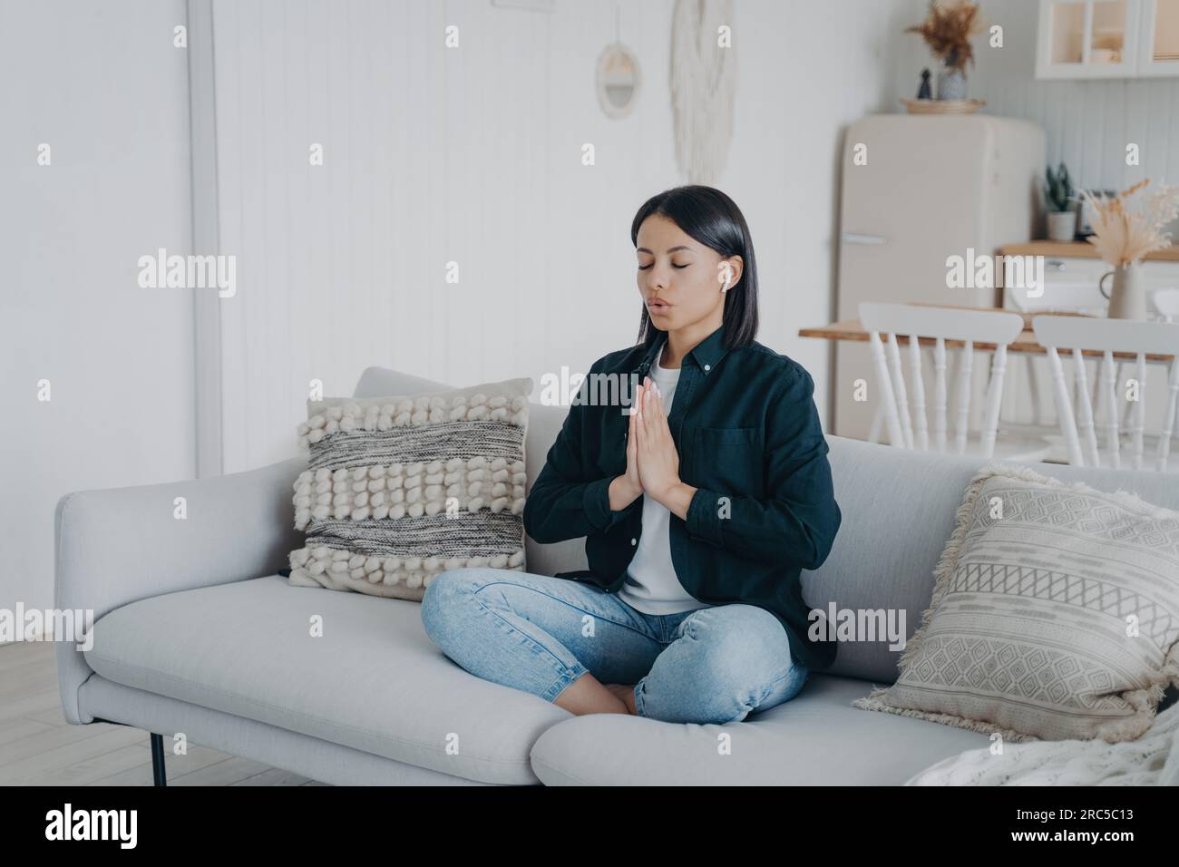 Ruhige Frau übt Yoga, meditiert in Lotus-Pose auf der Couch. Namaste-Geste, Entspannung zu Hause. Gesunder Lebensstil, Stressabbau. Stockfoto