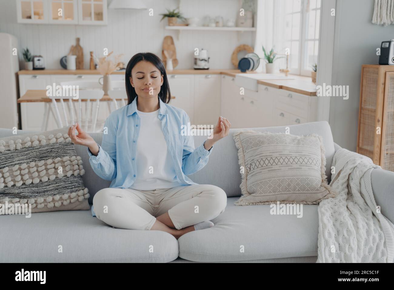 Eine heitere Frau übt Yoga, meditiert auf der Couch in Lotusposition. Atemübungen für mentale Balance, Angstlinderung. Gesunder Lebensstil zu Hause. Stockfoto