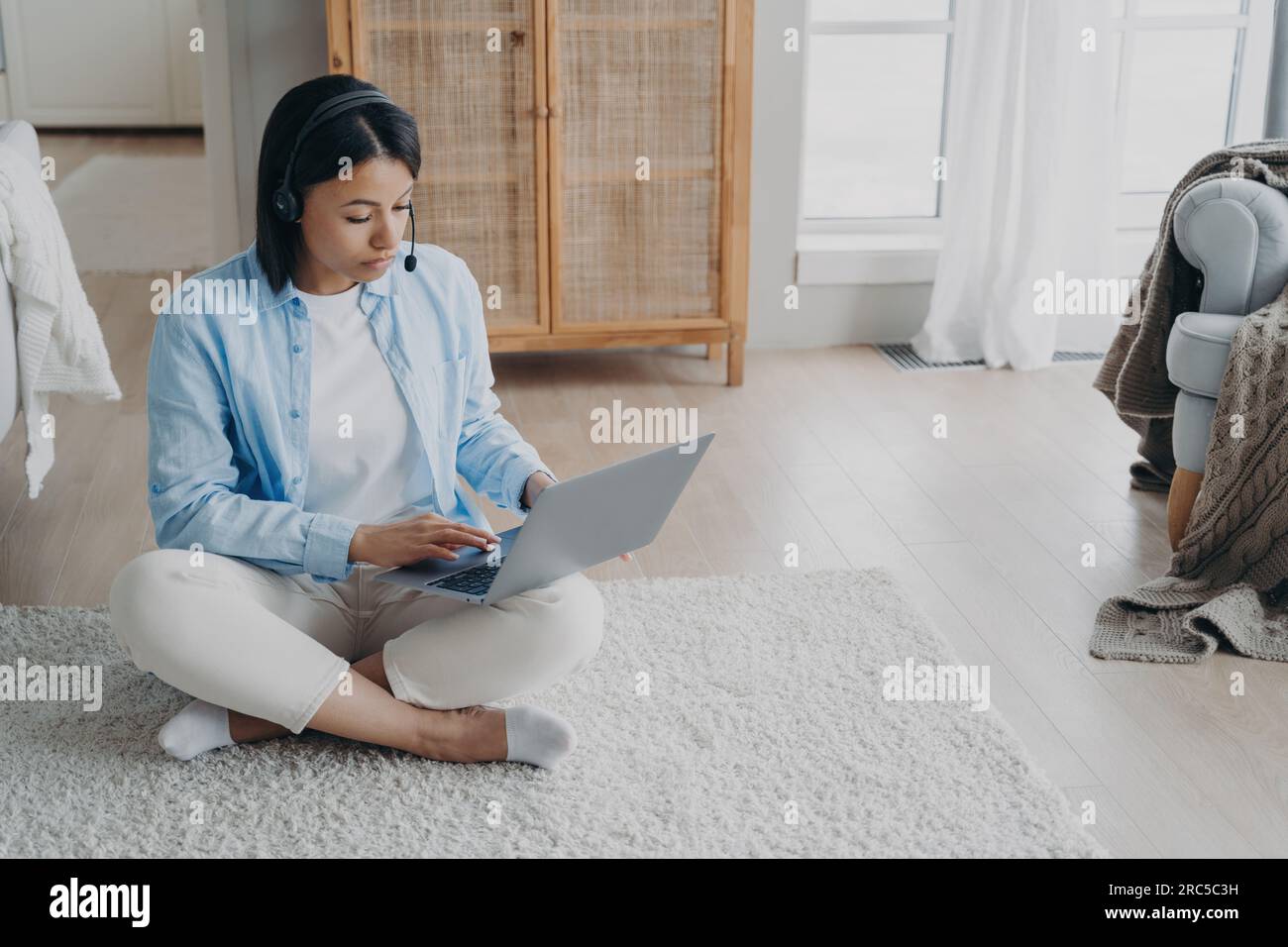 Fokussierter Freiberufler arbeitet auf einem Laptop, der zu Hause auf dem Boden sitzt. Seriöse Frau mit kabellosen Kopfhörern nimmt an einem informativen Webinar Teil und studiert online. E-Learning Stockfoto