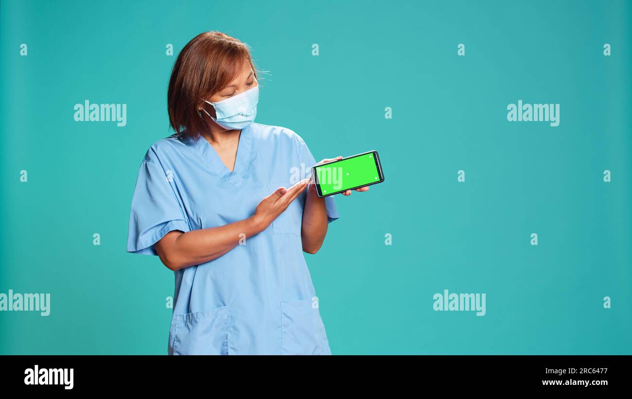 Krankenschwester präsentiert erläuterndes Gesundheitsband auf einem Mock-up-Chroma-Key-grünen Bildschirm. Krankenhausmitarbeiter hält das Telefon im Querformat, zeigt Video, isoliert im blauen Studiohintergrund Stockfoto