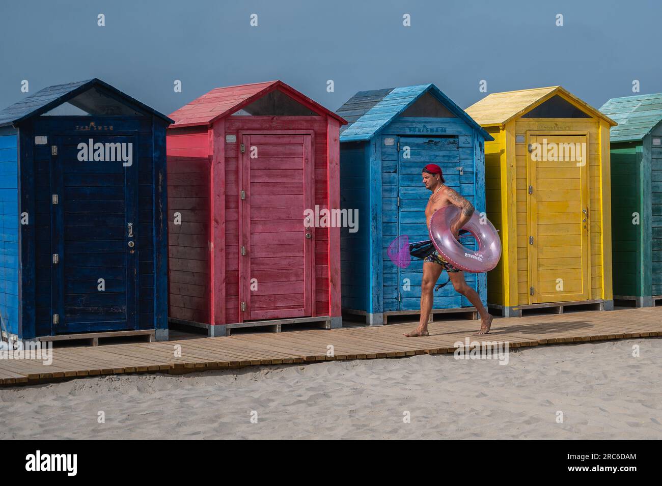 Alicante, Spanien. 12. Juli 2023. An einem heißen Sommertag am San Juan Beach führt ein Mann an einigen bunten Strandhütten vorbei, die einen aufblasbaren Schwimmer tragen. Die Staatliche Meteorologische Agentur (AEMET) hat rote Warnungen für hohe Temperaturen aktiviert, die durch eine Hitzewelle verursacht werden, die in einigen Gebieten Temperaturen über 40 Grad Celsius ansteigen wird. Kredit: Marcos del Mazo/Alamy Live News Stockfoto