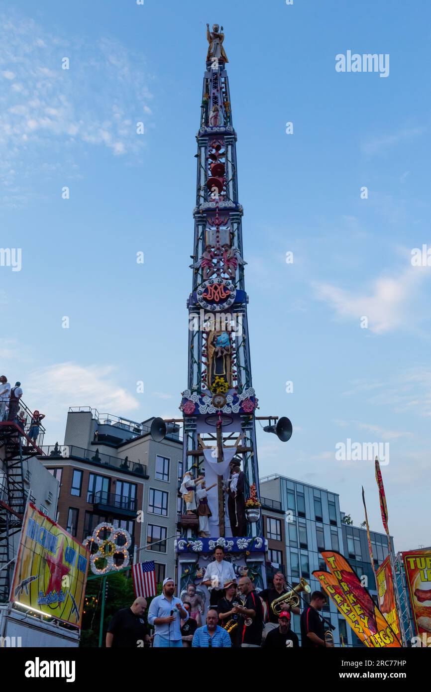 Brooklyn, New York, USA. 12. Juli 2023. Ein hoher Kirchturm mit Lilien und einer Band wird auf den Schultern von Gemeindemitgliedern getragen und im Rahmen des Fests unserer Lieben Frau vom Mount Carmel in Brooklyns Viertel Williamsburg durch die Gemeinde geführt. Der Kirchturm oder „giglio“ wird während des 11-tägigen Festes mehrmals auf der Straße hüpft oder „getanzt“, einschließlich dieses „Nachtlifts“. Während der giglio die Straße hinunterfährt, macht sich die Menge vor ihm frei. Kredit: Ed Lefkowicz/Alamy Live News Stockfoto