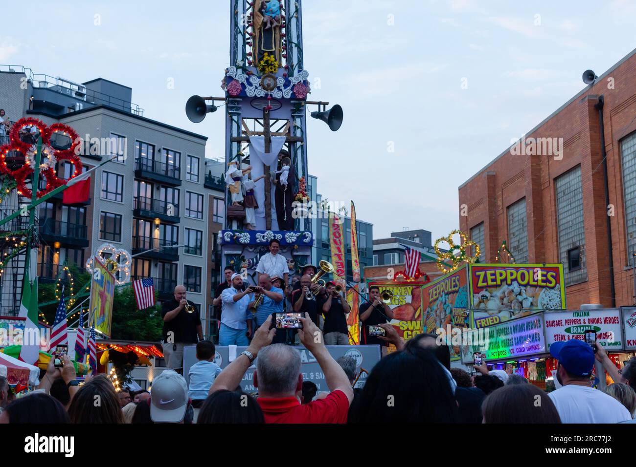 Brooklyn, New York, USA. 12. Juli 2023. Ein hoher Kirchturm mit Lilien und einer Band wird auf den Schultern von Gemeindemitgliedern getragen und im Rahmen des Fests unserer Lieben Frau vom Mount Carmel in Brooklyns Viertel Williamsburg durch die Gemeinde geführt. Der Kirchturm oder „giglio“ wird während des 11-tägigen Festes mehrmals auf der Straße hüpft oder „getanzt“, einschließlich dieses „Nachtlifts“. Während der giglio die Straße hinunterfährt, macht sich die Menge vor ihm frei. Kredit: Ed Lefkowicz/Alamy Live News Stockfoto