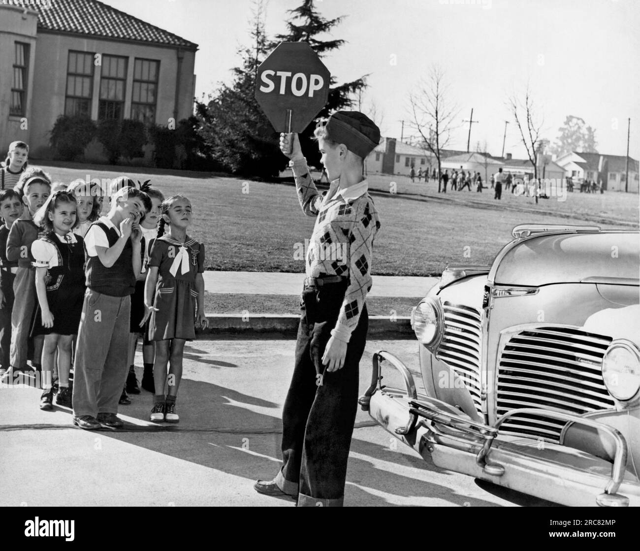 Vereinigte Staaten: c. 1948 Ein Schülerlotse hält sein Schild hoch, so dass die jüngeren Kinder die Straße nicht überqueren, bis er ihnen das Laufschild gegeben hat. Stockfoto