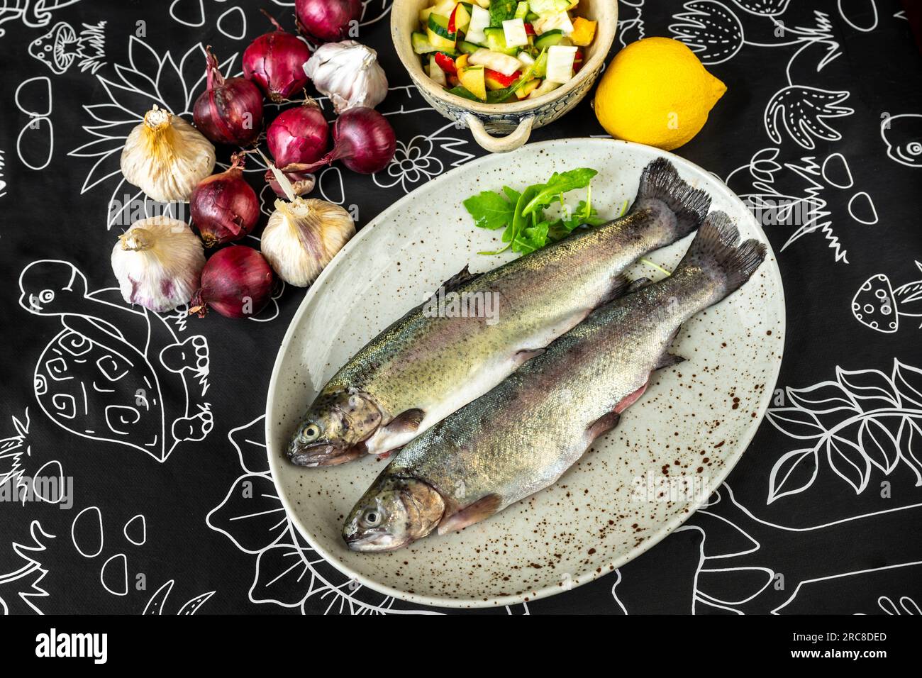 Zwei Regenbogenforellen auf dem Teller, viele Zwiebeln, Knoblauch und Zitrone auf schwarzem Tisch, Essenszubereitung. Stockfoto