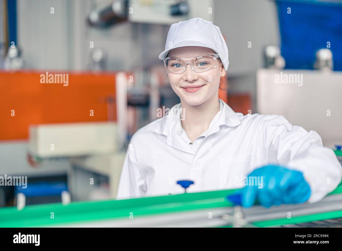 Glückliche junge weibliche Mitarbeiterin in der Förderbandproduktionslinie in der Lebensmittel- und Getränkefabrik mit weißer Hygienekleidung für Mitarbeiter Stockfoto