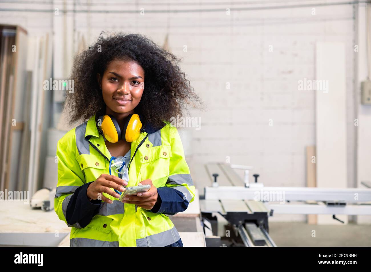 Porträt schwarze afrikanische Frau schlaue junge Ingenieurin mit Schutzausrüstung glücklich lächelnd mit Smartphone in der Holzmöbelfabrik Stockfoto
