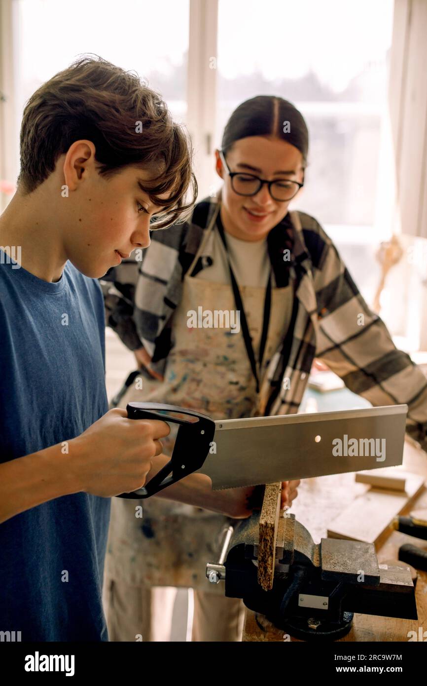 Männlicher Teenager-Schüler, der Holz schneidet, während er mit einer Lehrerin während des Tischlerkurses steht Stockfoto