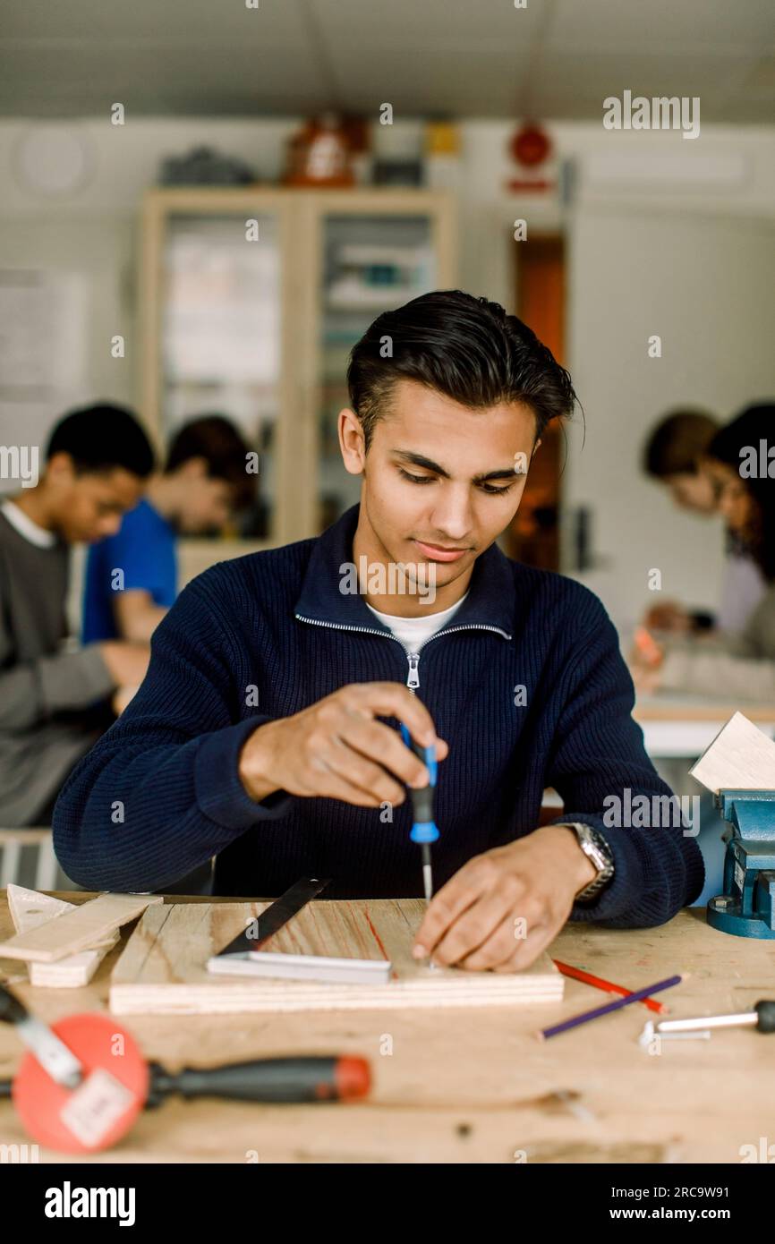 Männlicher Teenager-Schüler, der einen Schraubenzieher auf Holz benutzt, während er im Schreinerkurs in der Highschool lernt Stockfoto