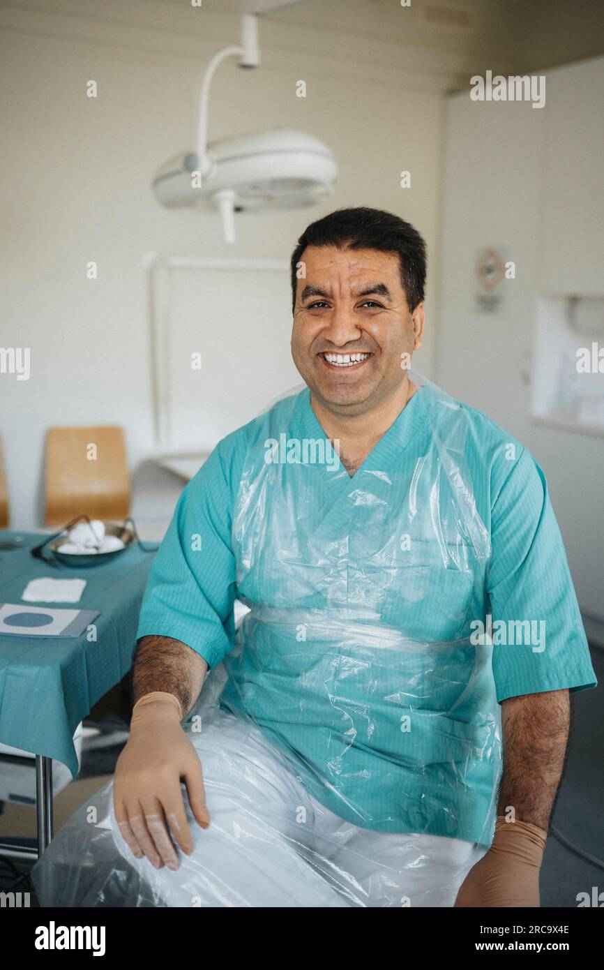 Porträt eines lächelnden männlichen Chirurgen, der schützende Sitzhaltung im ärztlichen Untersuchungsraum trägt Stockfoto
