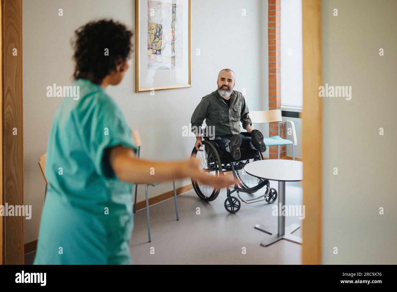 Ein Mann im Rollstuhl, der eine Krankenschwester ansieht, die sich an der Tür des Wartezimmers bewegt Stockfoto