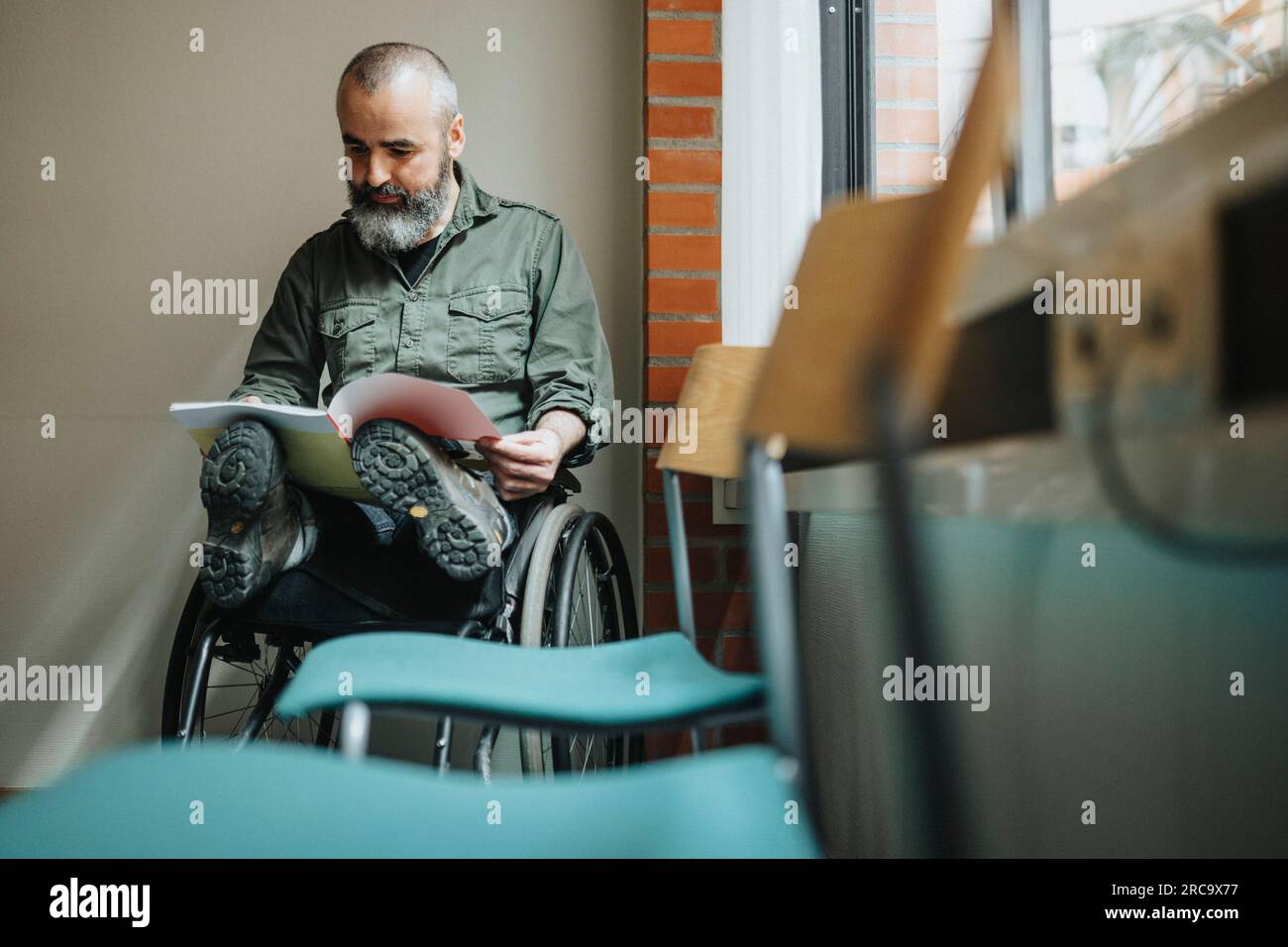 Reifer Mann mit Lesemagazin für Behinderte im Rollstuhl im Wartezimmer Stockfoto