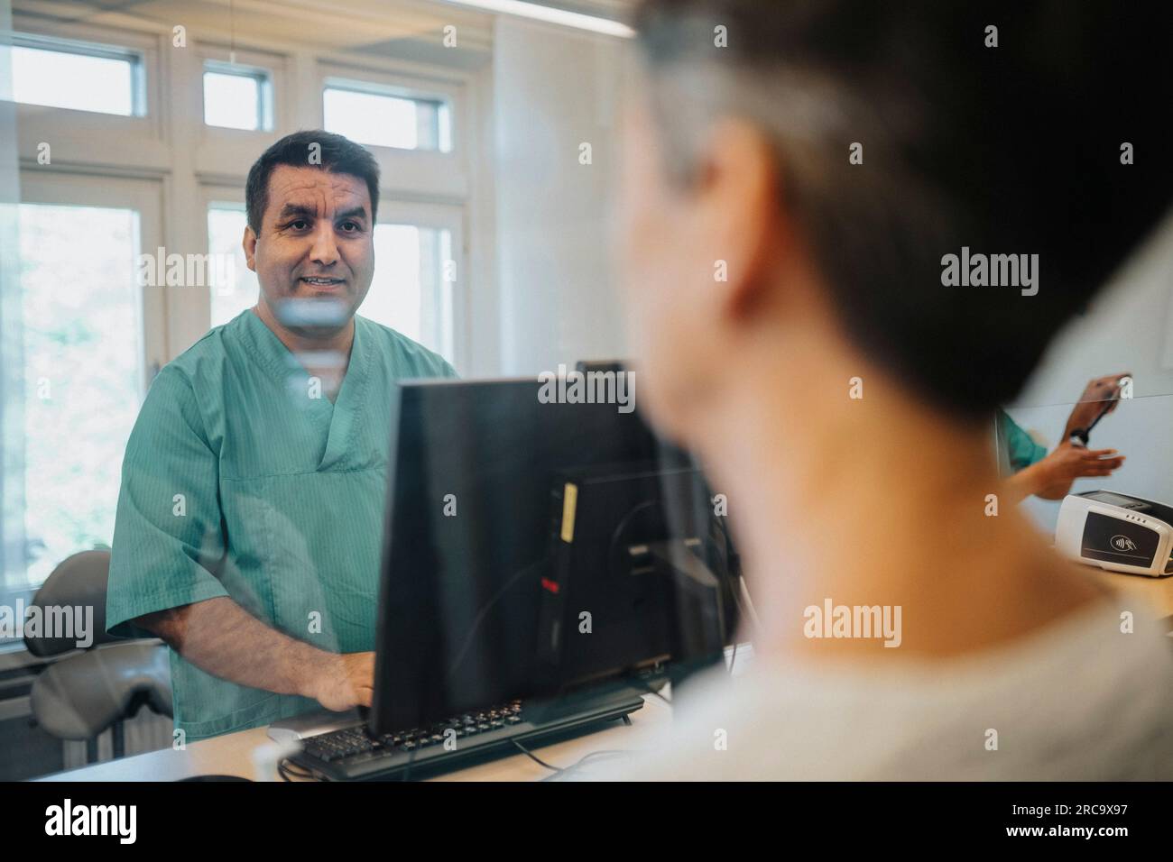 Männlicher Empfangsmitarbeiter, der mit dem Patienten durch Glaswand am Rezeptionstresen im Krankenhaus spricht Stockfoto