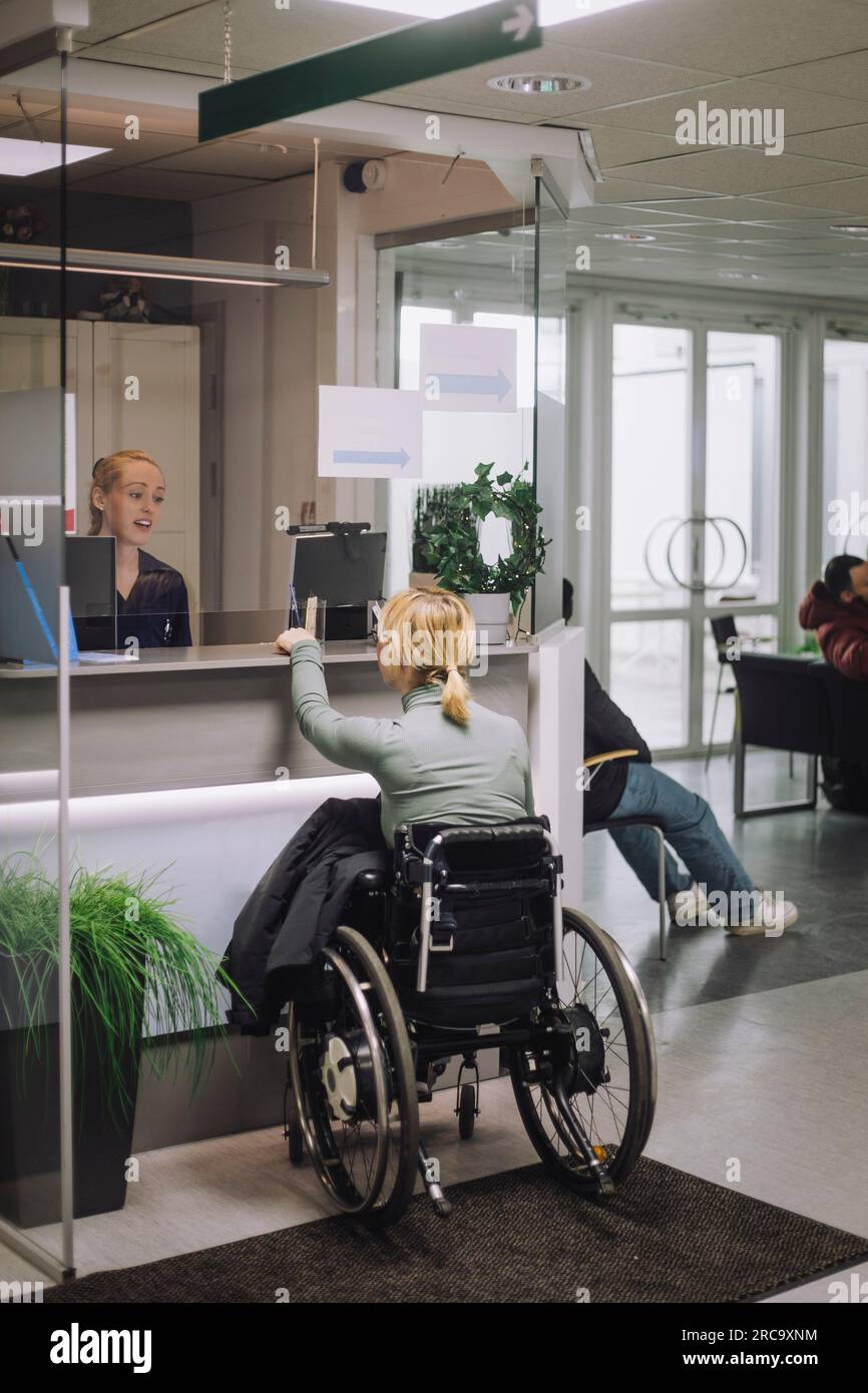 Weibliche Patientin sitzt auf Rollstuhl und erkundigt sich bei der Empfangsdame des Gesundheitszentrums Stockfoto