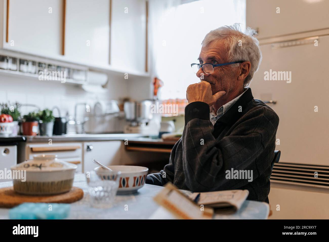 Rücksichtsvoller Senior-Mann mit Hand am Kinn, der zu Hause in der Küche sitzt Stockfoto