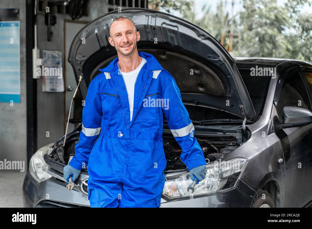 Fröhlicher, lächelnder Automechaniker in blauem Overall, der vor einem Auto steht und die Motorhaube öffnet Stockfoto