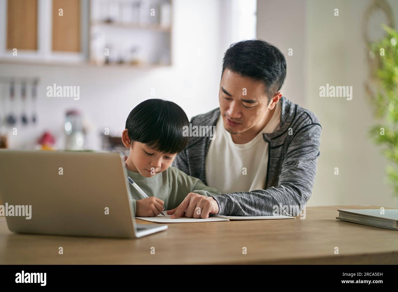 Junger asiatischer Vater, der an einem Tisch sitzt und seinen Sohn zu Hause unterrichtet Stockfoto