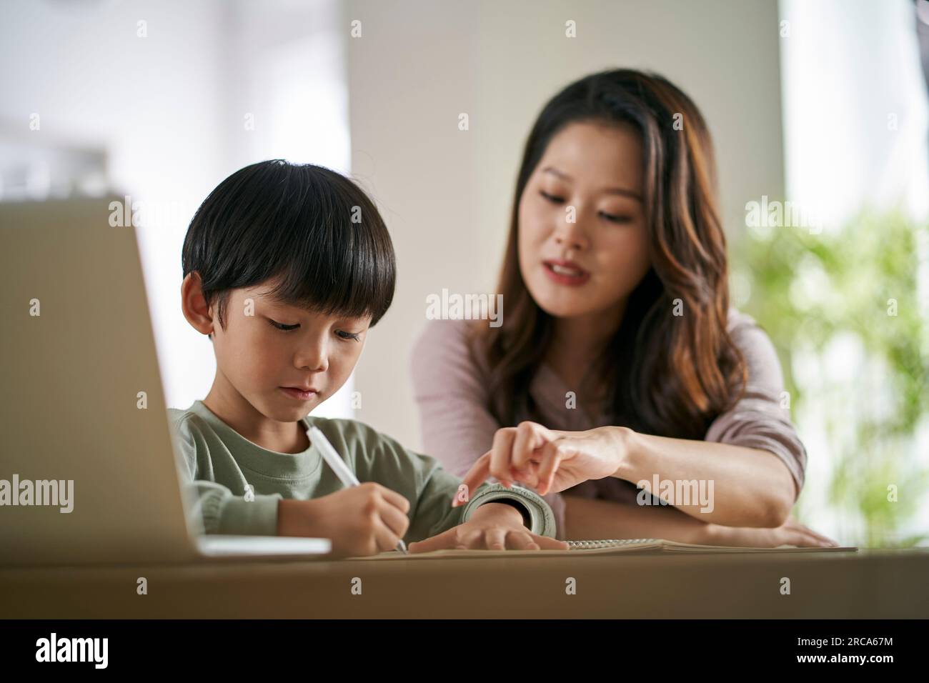Junge asiatische Mutter, die zu Hause am Tisch sitzt und dem Sohn beim Lernen hilft Stockfoto