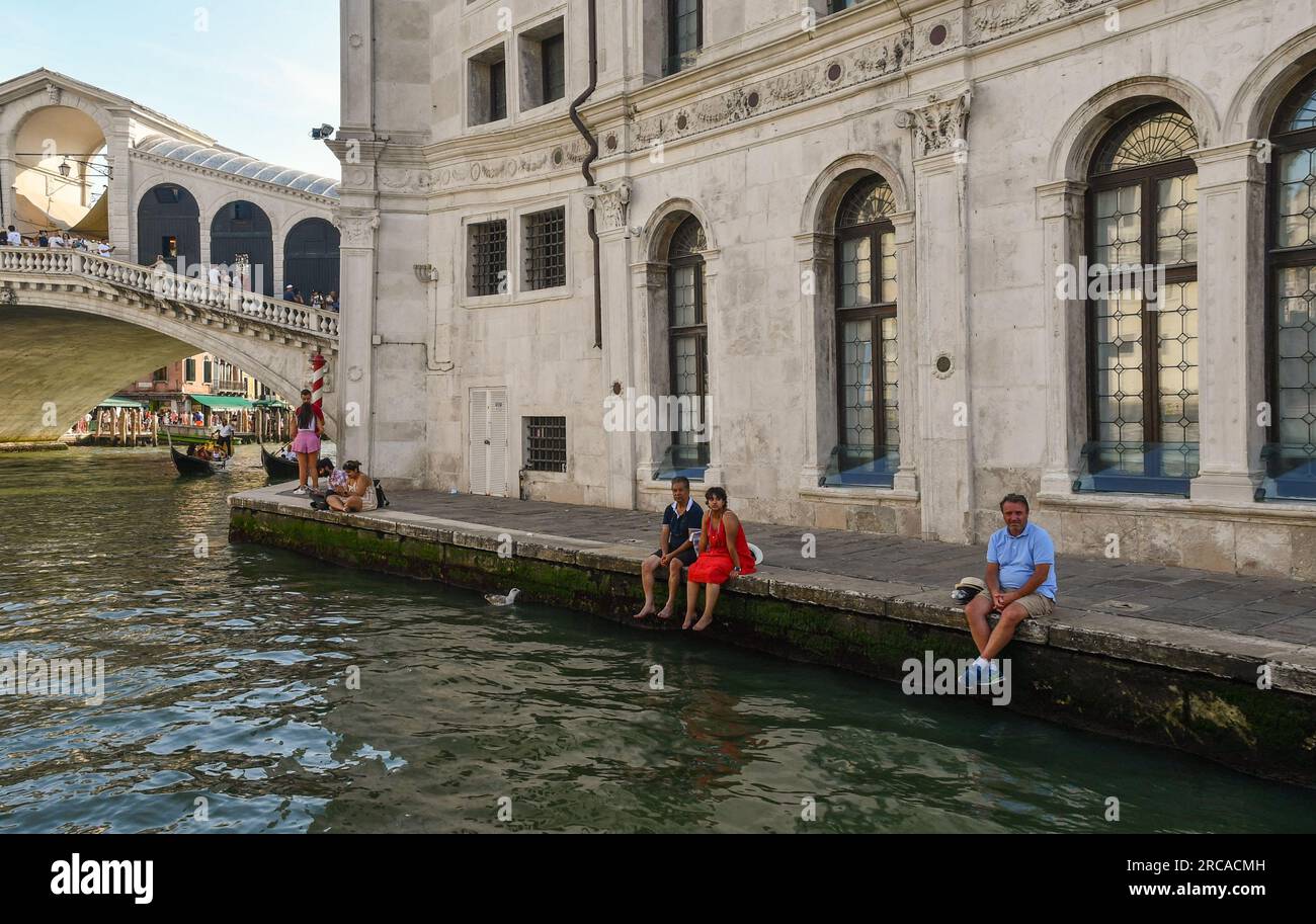 Touristen, die sich an einem heißen Sommertag vor dem Palazzo dei Camerlenghi auf dem Canale Grande mit der Rialtobrücke im Hintergrund in Venedig, Italien, ausruhen Stockfoto