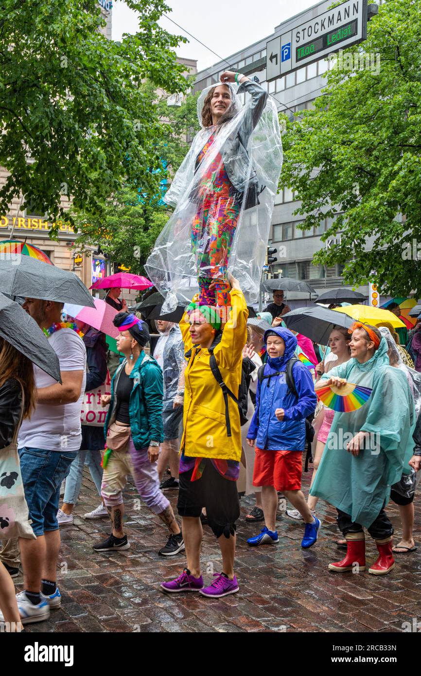 Frau im Einweg-Regenmantel steht auf den Schultern eines Freundes bei der Helsinki Pride 2023-Parade an einem Regentag in Helsinki, Finnland Stockfoto