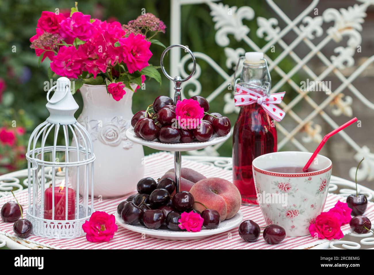 Tische mit frischen süßen Kirschen und Pfirsichen auf einem mehrstufigen Kuchenstand und Traubensaft in einer Tasse und einer Glasflasche Stockfoto