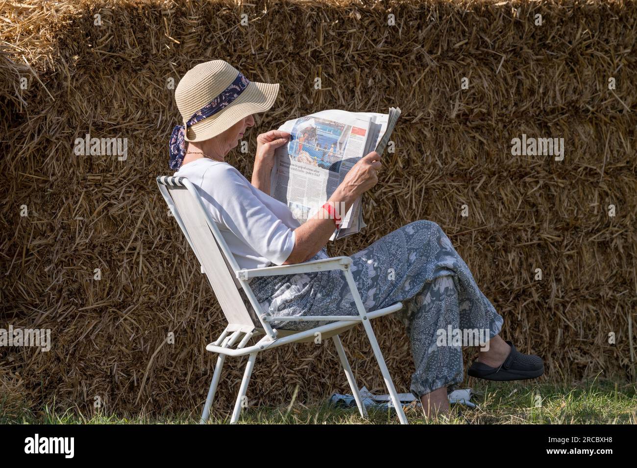 Ältere, weibliche, sitzende, eine Reihe Strohballen, die eine Zeitung lesen, während sie auf einem weißen metallenen Liegestuhl sitzen und einen Floppy-Hut tragen. Stockfoto