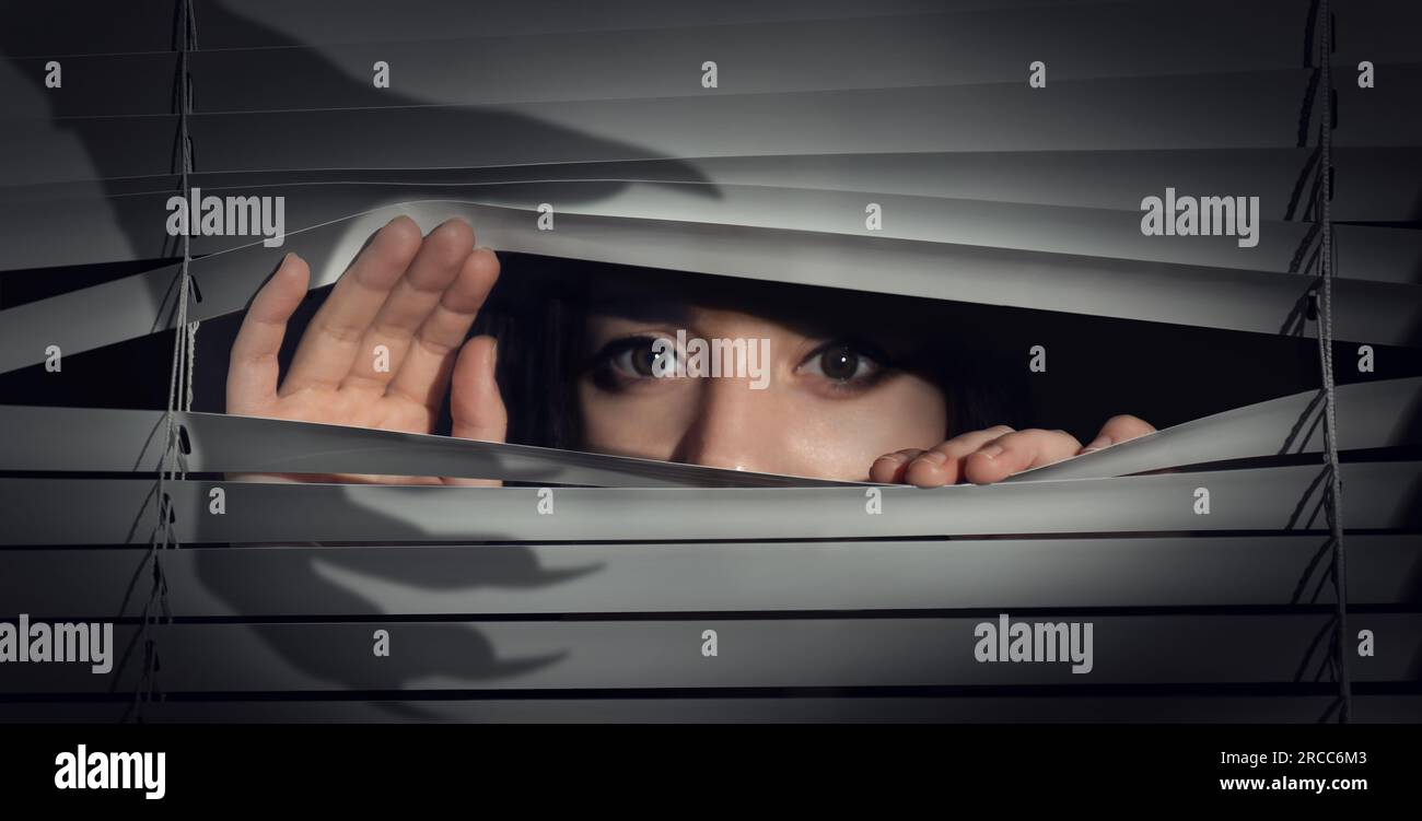 Besorgte Frau, die durch Fensterläden in die Dunkelheit schaut. Handschatten mit langen Klauen. Paranoia-Konzept Stockfoto