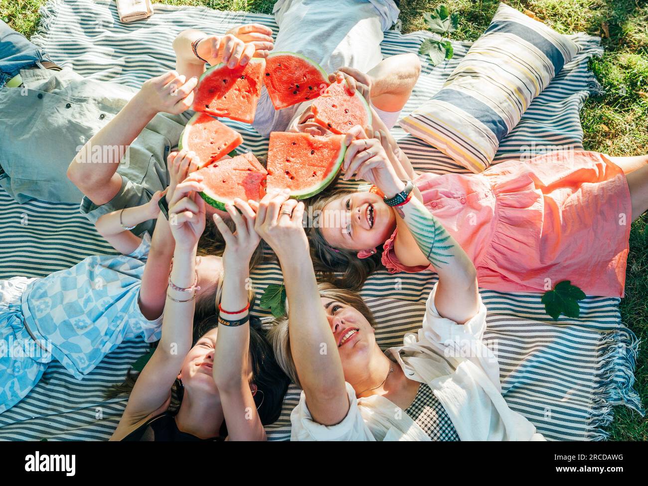 Junge Töchter mit Eltern, die an sonnigen Wochenendtagen auf einer Picknickdecke liegen, lächeln, lachen und rote, saftige Wassermelonen aufstellen. Familie Stockfoto