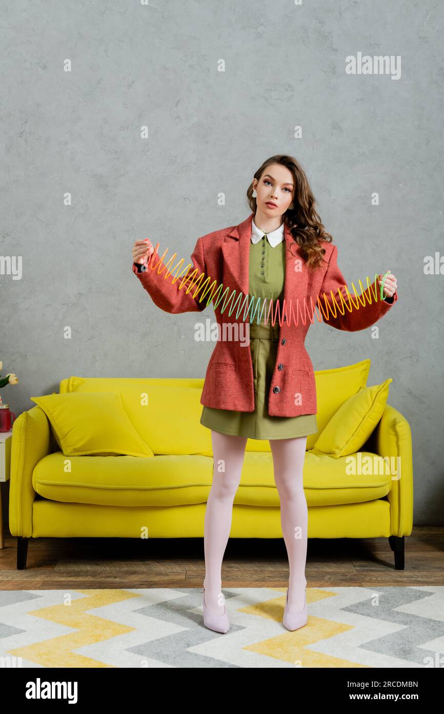 Junge brünette Frau, die sich wie eine Puppe ausgibt und mit Regenbogen-Slinky spielt, in die Kamera schaut, modernes Wohnzimmer mit gelbem Sofa, kindisch, altmodisch, n Stockfoto