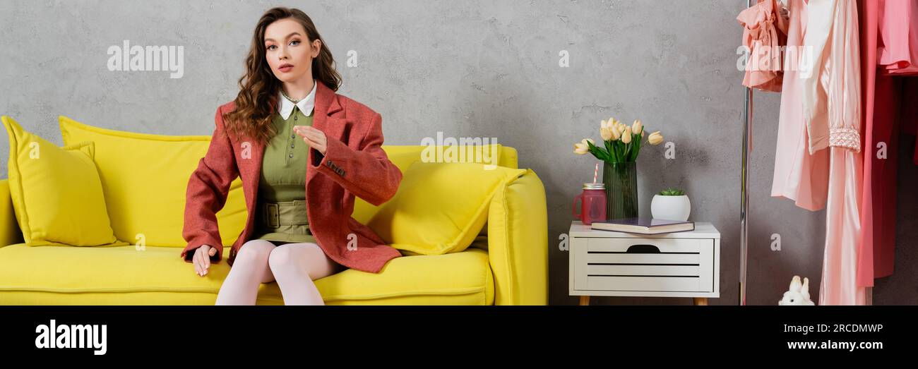 Konzeptfotografie, Posen wie eine Puppe, junge Frau mit welligen Haaren, die auf der gelben Couch sitzt, Gesten, stilvolle Inneneinrichtung, Vase mit Tulpen, Look Stockfoto