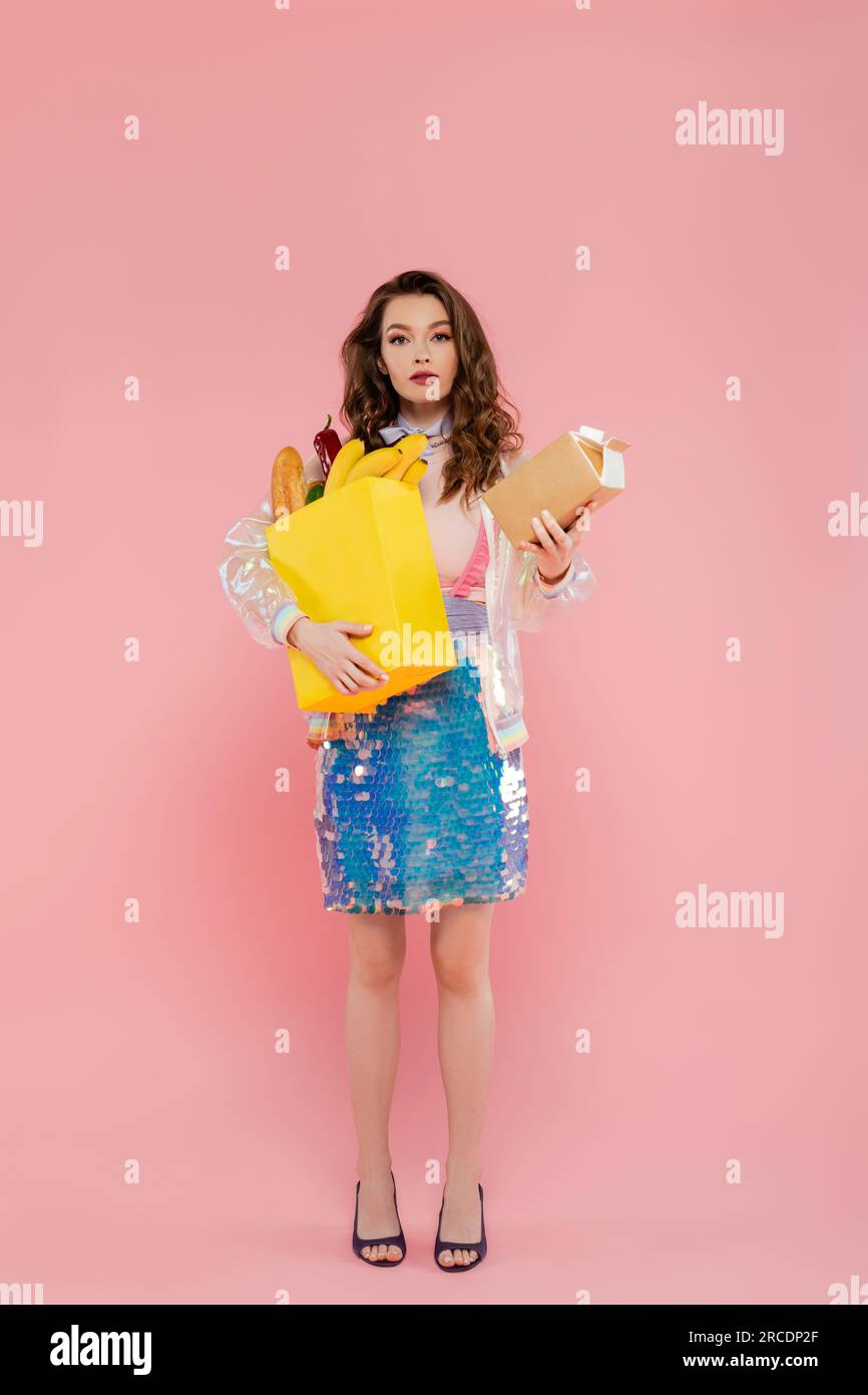Hausfrauenkonzept, attraktive junge Frau mit Einkaufstasche und Karton mit Milch, Modell mit welligem Haar auf pinkfarbenem Hintergrund, konzeptionelle Fotografie, Stockfoto