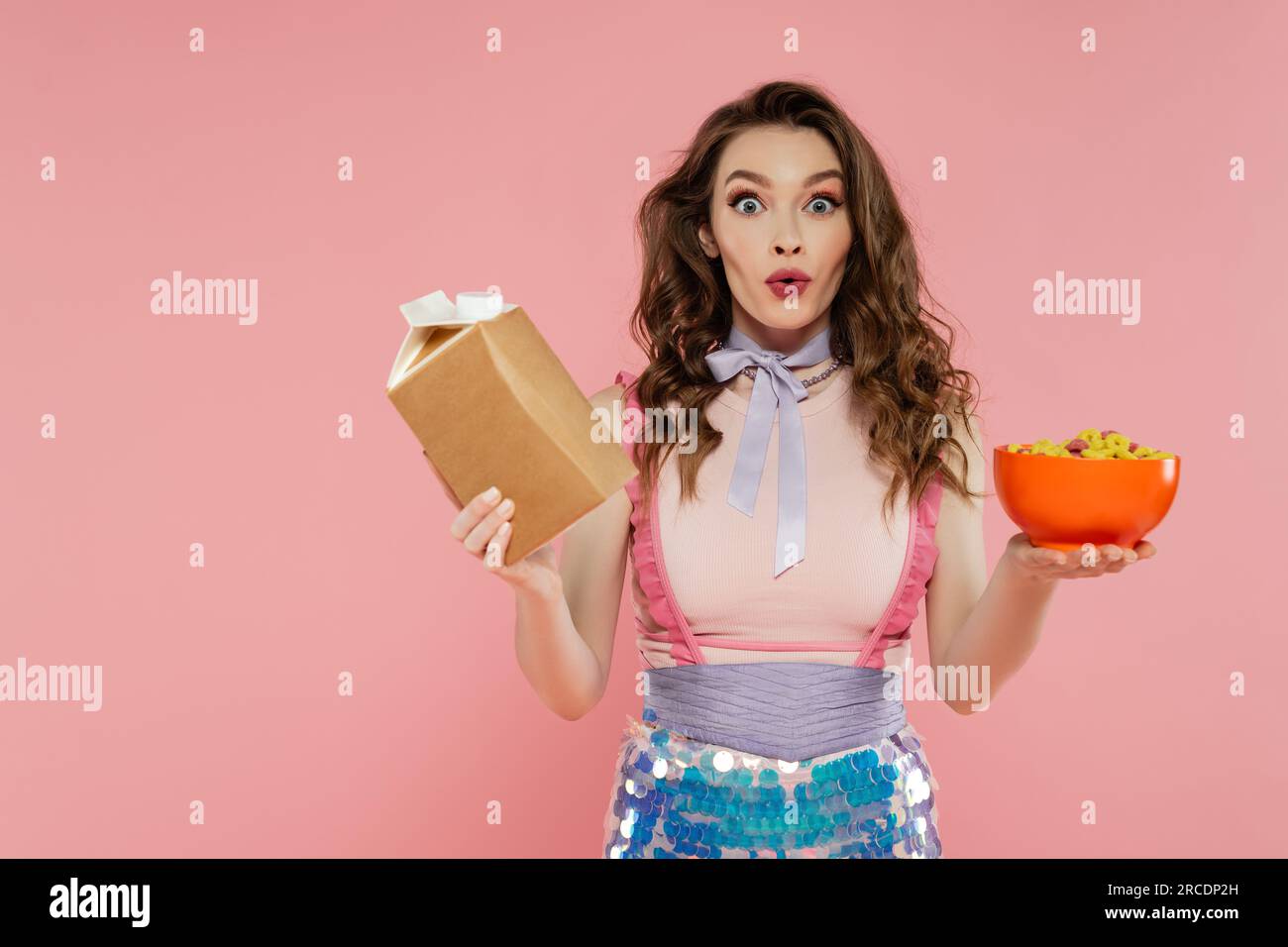 Hausfrauenkonzept, schockierte junge Frau, die sich wie eine Puppe ausgibt, mit Essen zum Mitnehmen und einer Schüssel mit Cornflakes, rosa Hintergrund, Konzeptfotografie, h Stockfoto