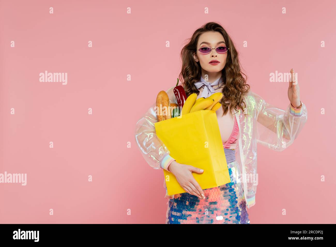 Hausfrauenkonzept, junge Frau in Sonnenbrille trägt Papiertasche mit Lebensmitteln, braunes Modell mit welligem Haar, das sich wie eine Puppe auf pinkfarbenem Hintergrund ausstellt, c Stockfoto
