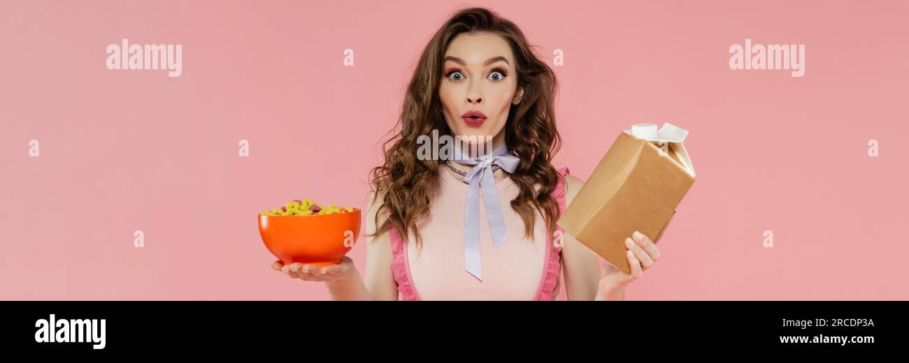 Hausfrauenkonzept, schockierte junge Frau, die sich wie eine Puppe ausgibt, mit Essen zum Mitnehmen und einer Schüssel mit Cornflakes, rosa Hintergrund, Konzeptfotografie, h Stockfoto