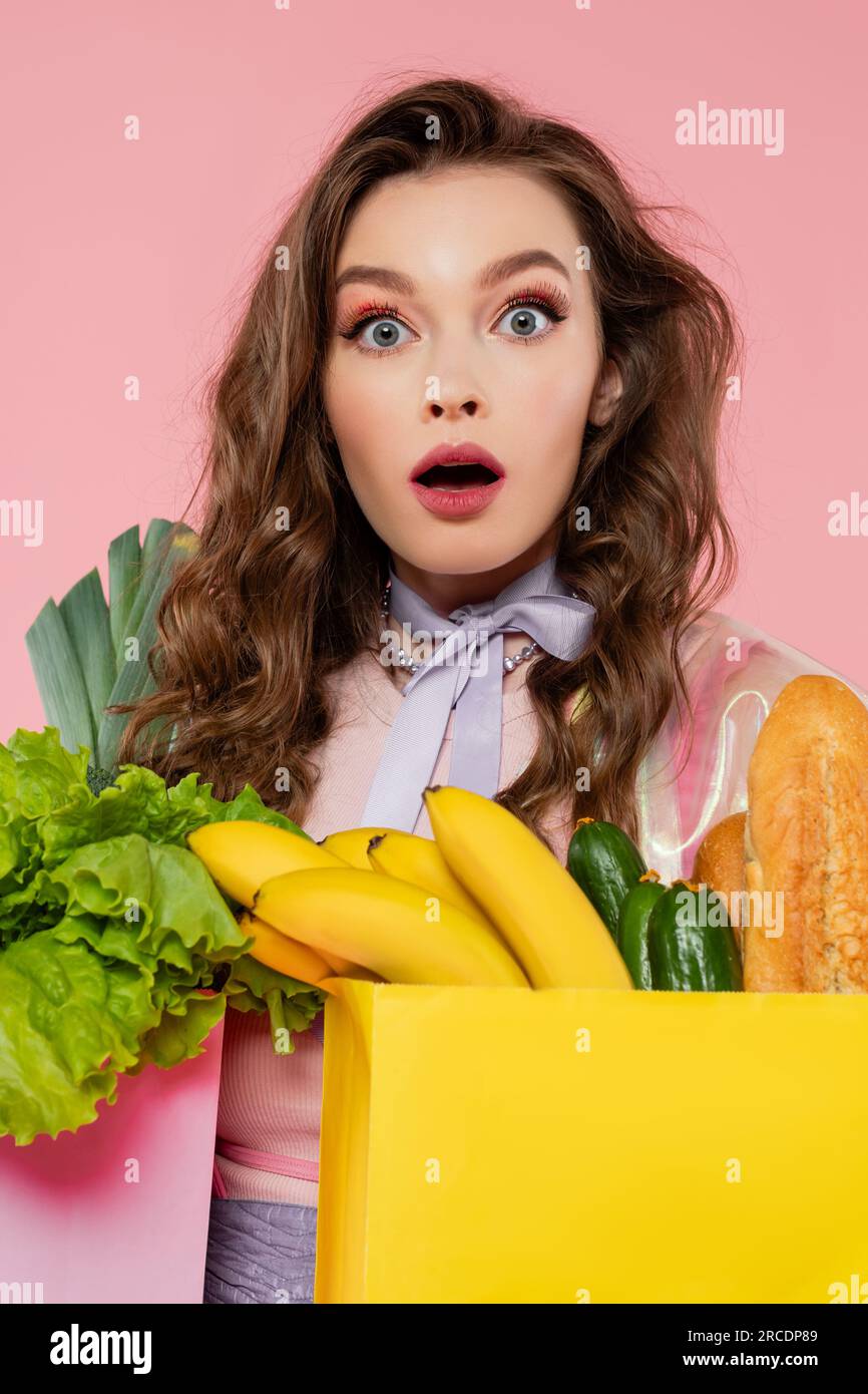 Hausfrauenkonzept, schockierte Frau, die Einkaufstüten mit Gemüse und Bananen trägt, Modell mit welligem Haar auf pinkfarbenem Hintergrund, Konzeptfotografie Stockfoto
