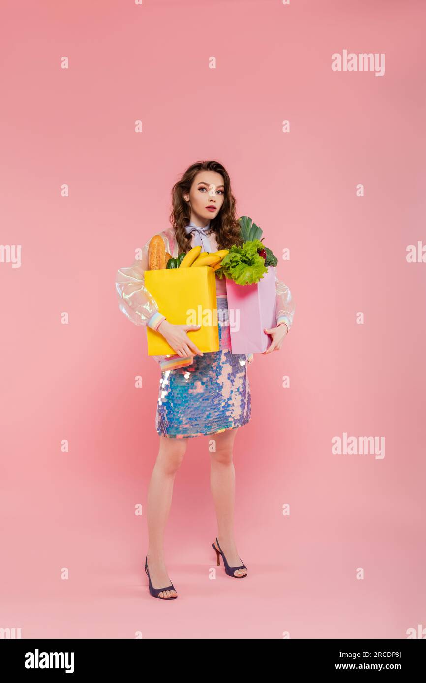 Hausfrauendienst, attraktive junge Frau, die Papiertaschen mit Lebensmitteln trägt, Modell mit welligen Haaren auf pinkfarbenem Hintergrund, Konzeptfotografie, Stockfoto