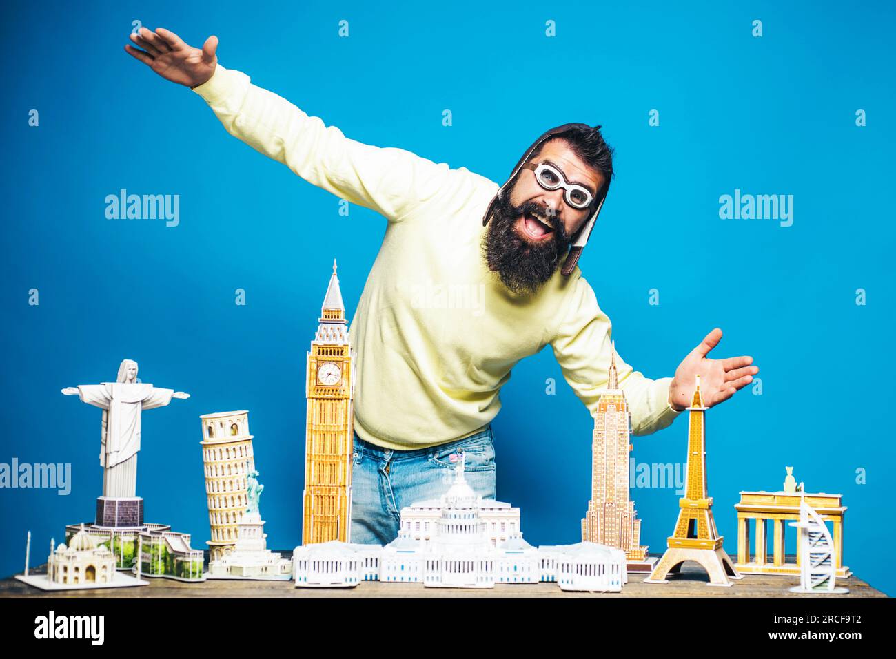 Ein glücklicher bärtiger Mann mit Pilotenhelm und Brille glaubt, über den Tisch fliegen zu können, mit Miniaturbauten Modelle, die aus 3D Puzzles bestehen. Weltarchitektur Stockfoto