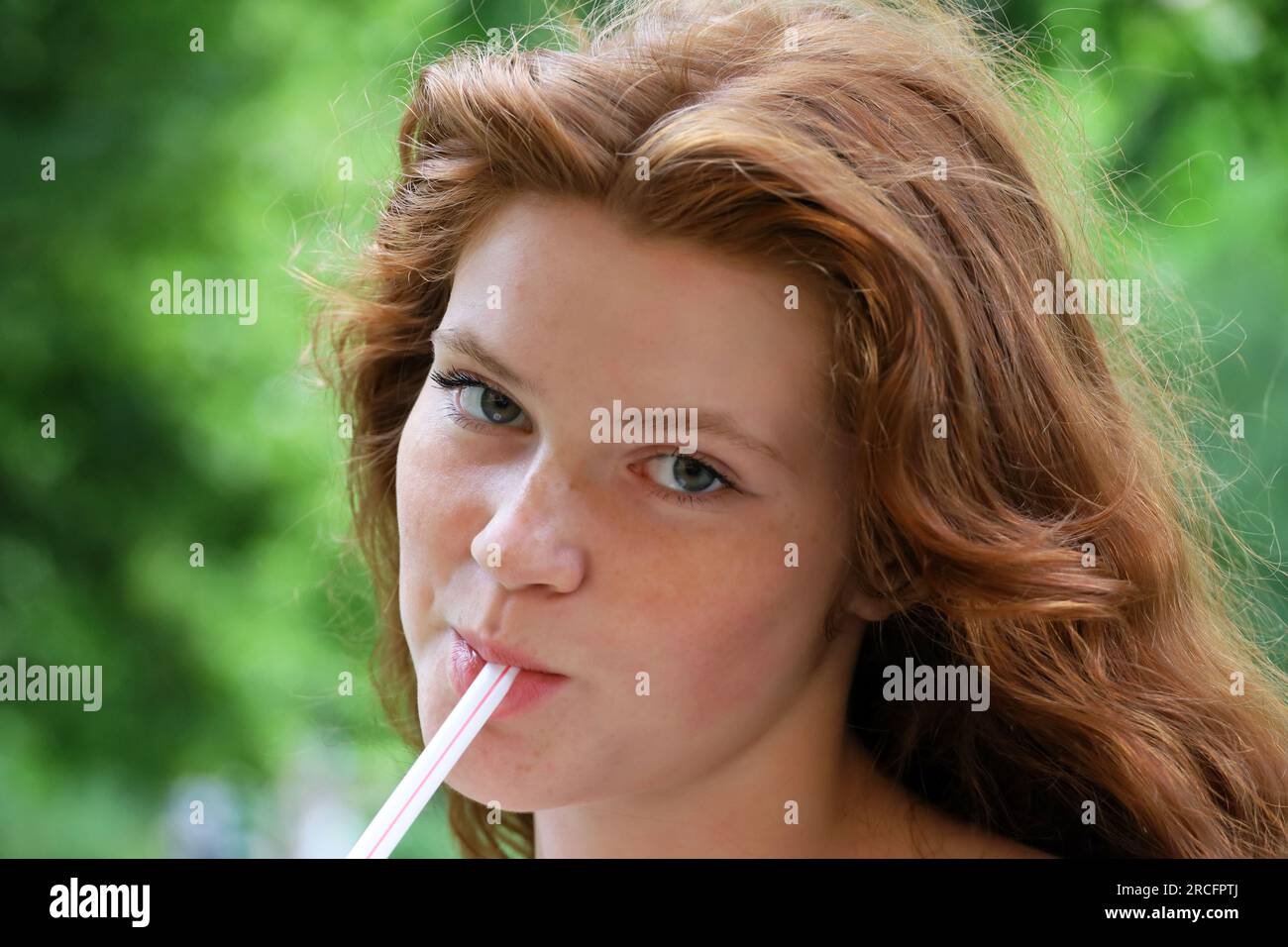 Portrait eines hübschen Ingwermädchens mit langen roten Haaren und Sommersprossen, das im Sommerpark einen Cocktail mit Strohhalm trinkt. Ein junges sinnliches Modell Stockfoto