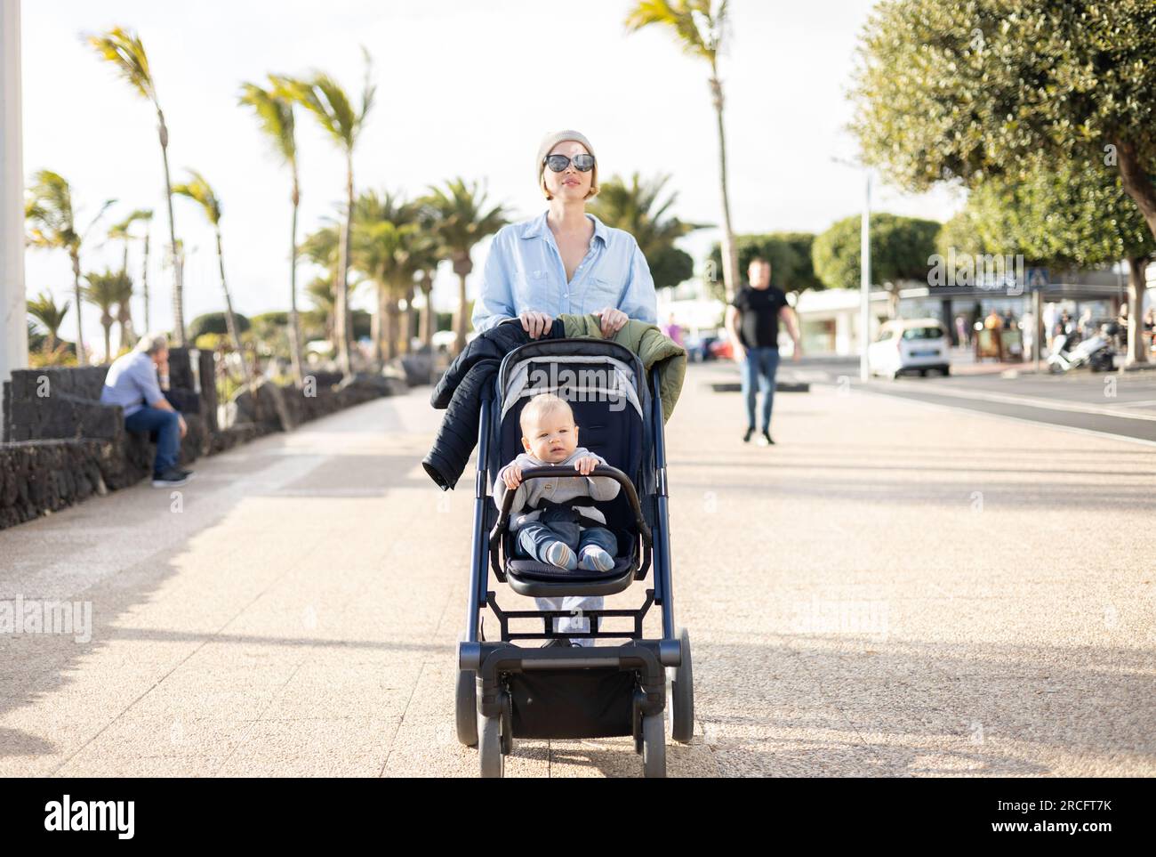 Eine Fashible Mutter, die ihr kleines Kind im Kinderwagen entlang der palmengesäumten Strandpromenade von Puerto del Carmen, Lanzarote, kanarienvogel i schubst Stockfoto