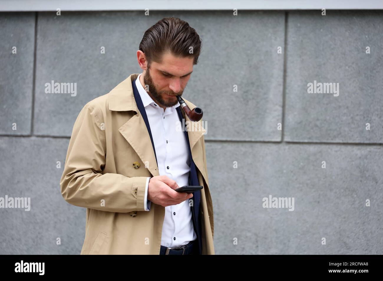 Ein Mann mit einer rauchenden Pfeife, der einen Regenmantel trägt und auf den Smartphone-Bildschirm schaut, während er auf einer Straße steht Stockfoto