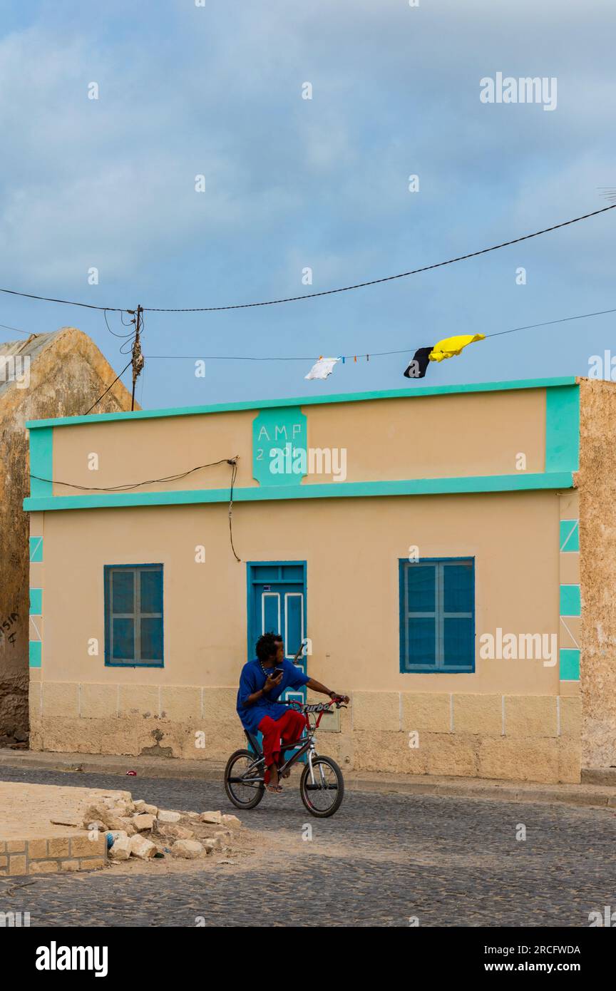 Ein afrikanischer Mann, der an einem Haus vorbeiradelt, während er auf seinem Handy, Boa Vista, Kap Verde, Afrika sitzt Stockfoto