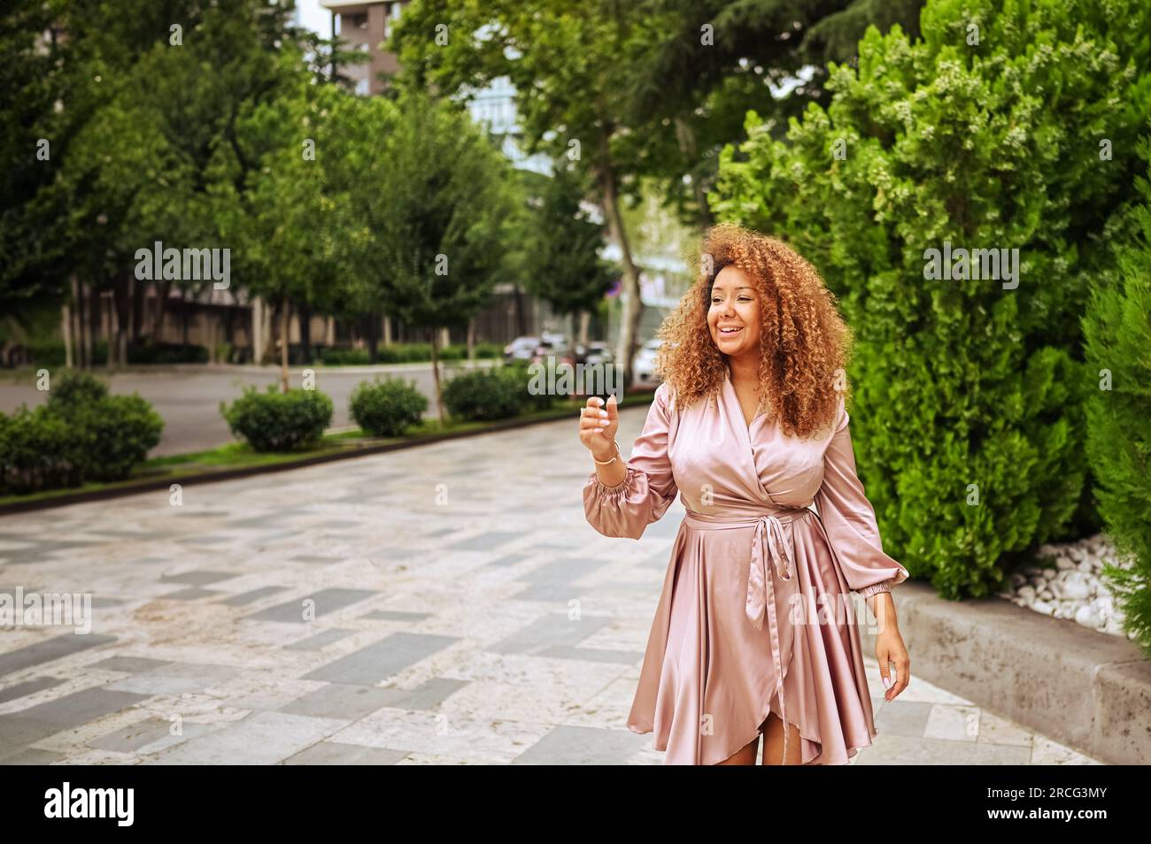 Eine junge, wunderschöne afroamerikanische Frau macht einen Spaziergang im öffentlichen Sommerpark. Eine glückliche Frau in Übergröße, die ein seidenrosa Kleid trägt, lächelt durch die Straßen der Stadt Stockfoto