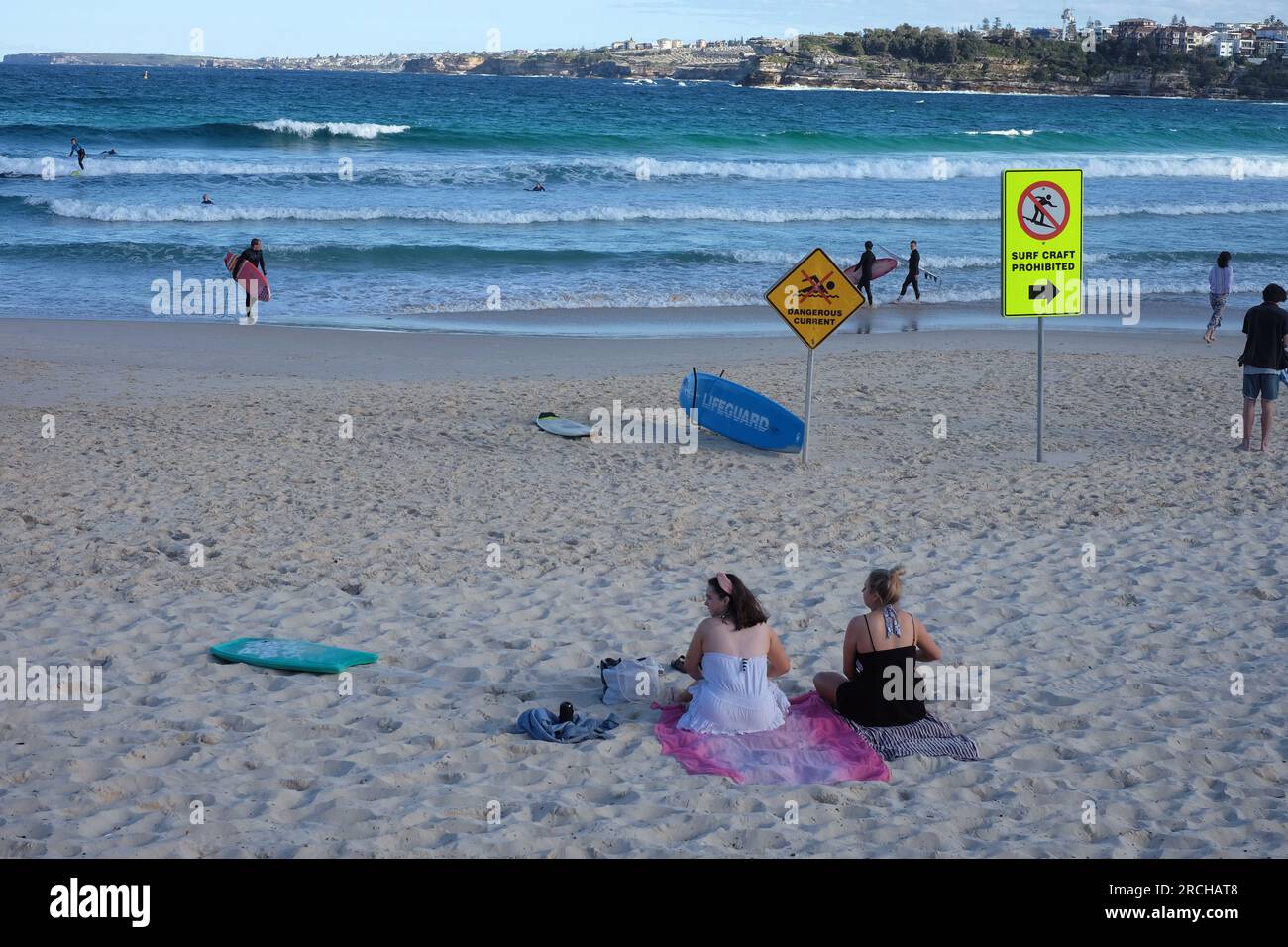 North Bondi, zwei Frauen in Sommerkleider, die an einem fast leeren Strand sitzen und die Brandung beobachten, zwei Schilder lesen, gefährliche Strömung, Surfboote verboten Stockfoto