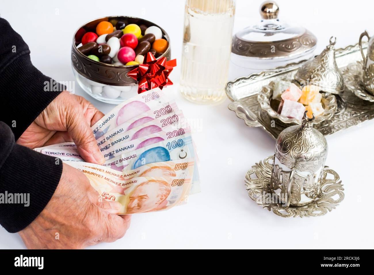 Türkische Banknoten in der Hand eines älteren Menschen auf weißer Oberfläche mit köln, bunten Süßigkeiten und türkischem Kaffeeset. Festmahlbonus spiegelt sich in Pensionen wider Stockfoto