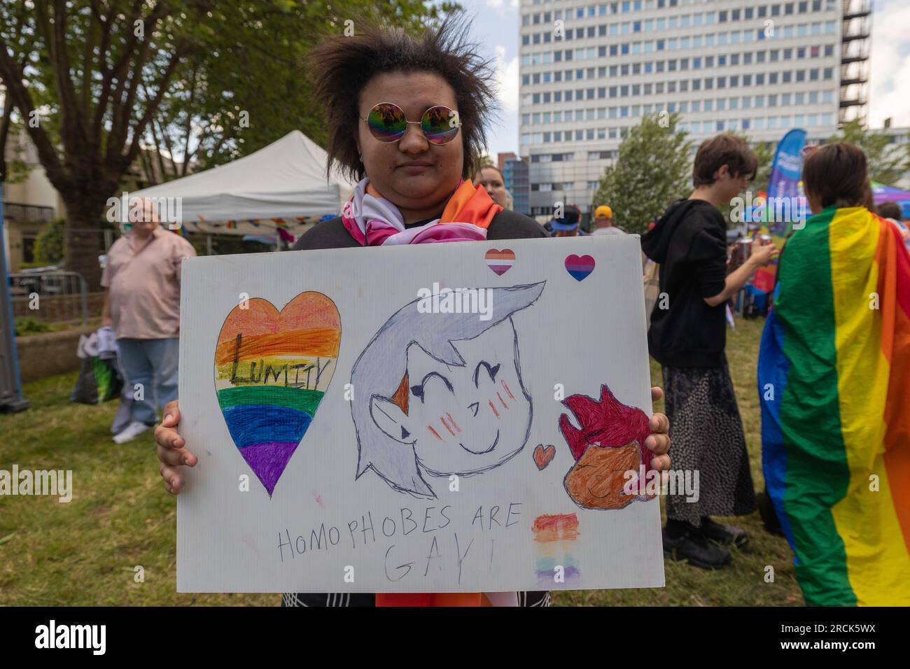 Southend on Sea, Großbritannien. Juli 2023. Ein handgefertigtes LGBTQ+-Stolz-Zeichen, das von einem Teilnehmer bei Southend Pride in Southend-on-Sea, Essex, gehalten wird. Auf dem Schild befinden sich Kunstwerke im Cartoon-Stil, Regenbogenherzen und der Satz „Homophobe are Gay!“ In Regenbogenschriftzug. Die Person trägt Sonnenbrille und Schal in Regenbogenfarben, umgeben von anderen Besuchern in einem Park mit Bürgerhäusern im Hintergrund. Tausende von Menschen nehmen an der jährlichen Pride Parade on the High Street und dem Festival auf dem Warrior Square teil. Penelope Barritt/Alamy Live News Stockfoto