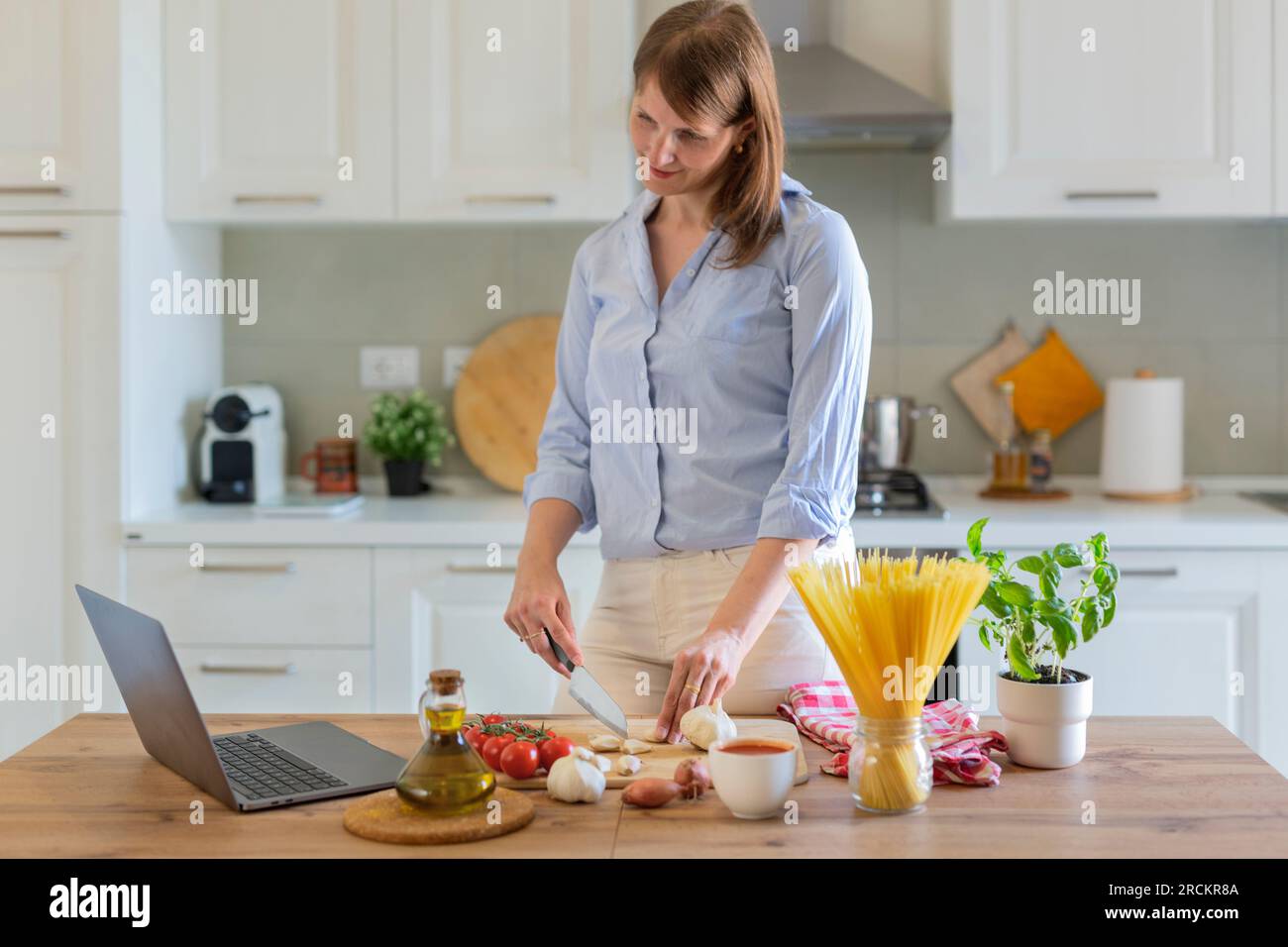 Eine junge Frau kocht zu Hause in der Küche, sie sieht sich ein Videorezept auf einem Laptop an und arbeitet mit dem Teig auf dem Tisch, sie schneidet Knoblauch. Italienischer cui Stockfoto