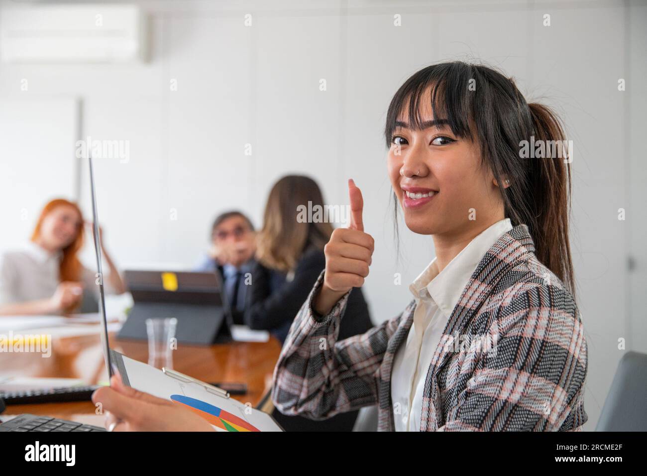 Eine Geschäftsfrau mit erhobenen Daumen im Büro bei der Arbeit, mehrere ethnische Gruppen Stockfoto