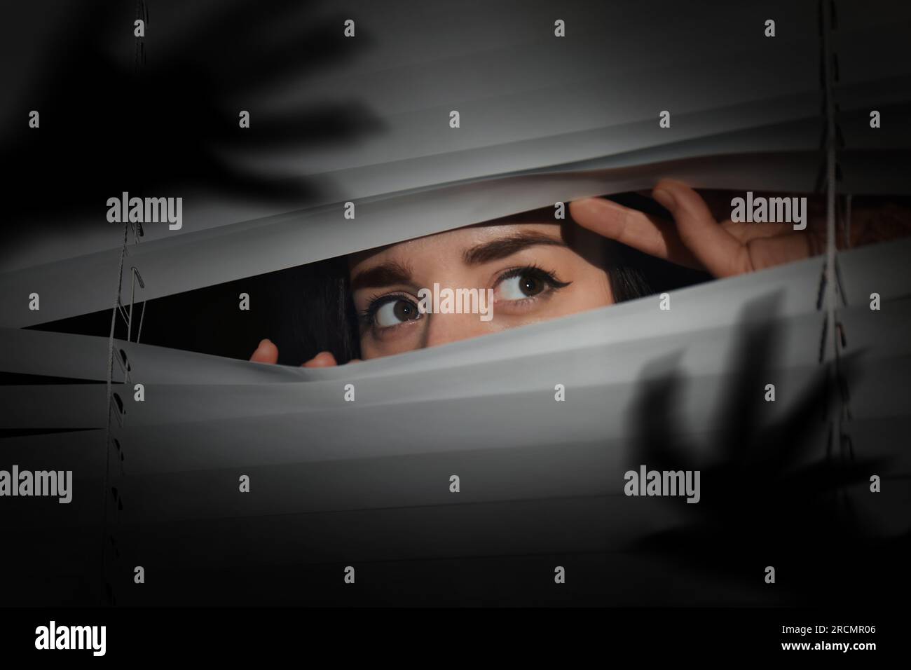 Besorgte Frau, die durch Fensterläden in die Dunkelheit schaut. Handschatten mit langen Klauen. Paranoia-Konzept Stockfoto