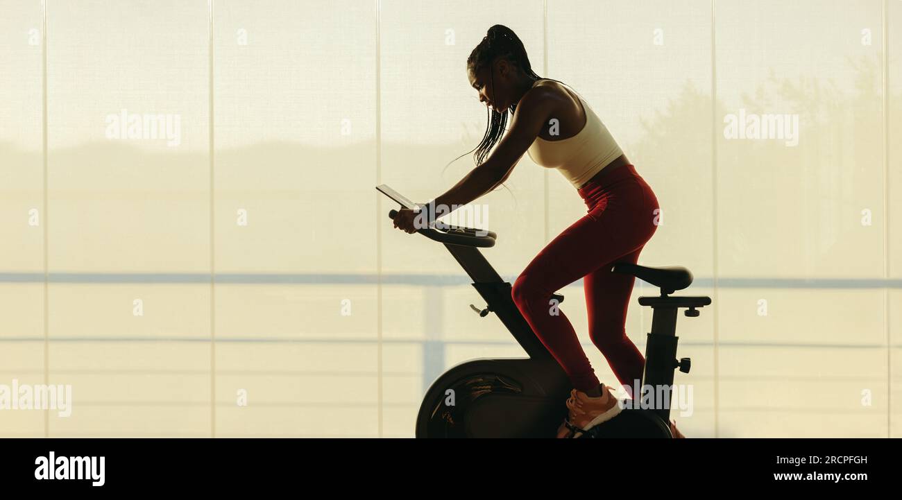 Junge schwarze Frau, die zu Hause in einem Online-Fitnesskurs trainiert. Sportliche Frau, die auf einem Sportmotorrad in die Pedale tritt und ihr Ja auf dem digitalen Tablet A fixiert ist Stockfoto
